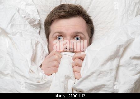 Nightmare. Caucasian man is scared, he hides under the covers and looks with big eyes, lying in bed. Psychological health concept. Stock Photo
