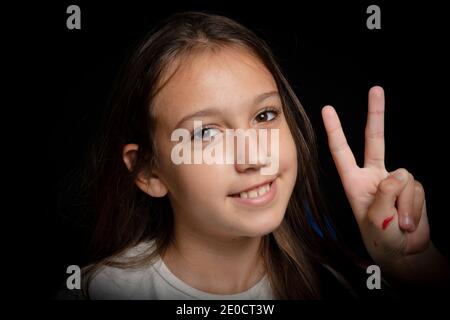 Close up portrait of a cheerful little girl making the victory gesture with her hand stained in red paint on black studio background. Joy concept Stock Photo