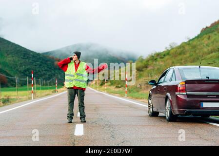 Female driver in reflective vest standing on road near broken car in mountainous area and talking on smartphone while calling for help Stock Photo