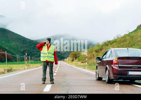 Female driver in reflective vest standing on road near broken car in mountainous area and talking on smartphone while calling for help Stock Photo