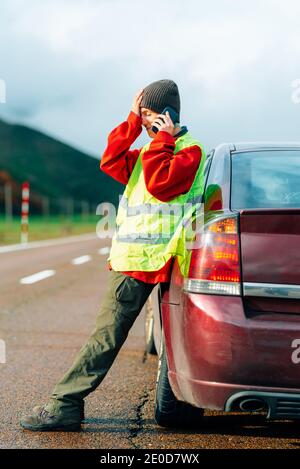 Side view of bewildered female driver in reflective vest leaning on broken car while standing on road in highlands and calling insurance for help Stock Photo