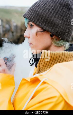 Side view of relaxed female in outerwear smoking vape and exhaling cloud of smoke while standing on Cobijero Beach and looking away Stock Photo