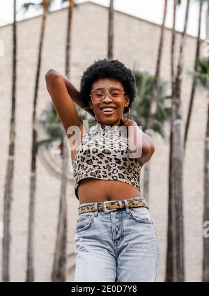 From below of positive young curly haired African American female in casual outfit and trendy sunglasses standing against blurred building on street Stock Photo