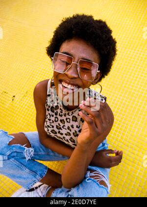 From above of optimistic young African American female in trendy ripped jeans and stylish sunglasses sitting on yellow floor and laughing happily with Stock Photo