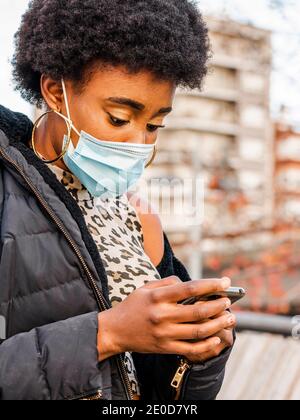 Modern young black female with Afro hairstyle wearing warm jacket and protective mask browsing mobile phone while standing near city building Stock Photo