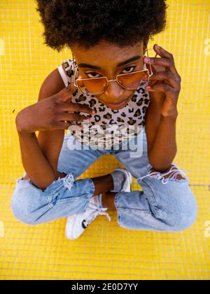 From above young African American female in trendy ripped jeans and stylish sunglasses sitting on yellow floor Stock Photo