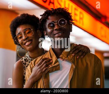 Happy young African American man and Woman with curly hair wearing stylish sunglasses looking at camera while standing together near urban building Stock Photo