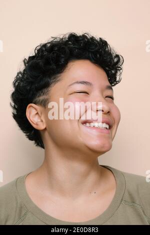 Portrait of young Latina Woman with closed Asian eyes smiling, isolated vertical photo, beige background Stock Photo