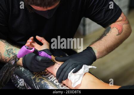 Unrecognizable concentrate bearded male master in black face mask and gloves creating tattoo on arm of crop faceless client in salon Stock Photo