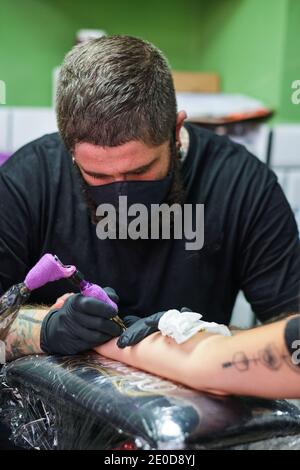 Unrecognizable concentrate bearded male master in black face mask and gloves creating tattoo on arm of crop faceless client in salon Stock Photo