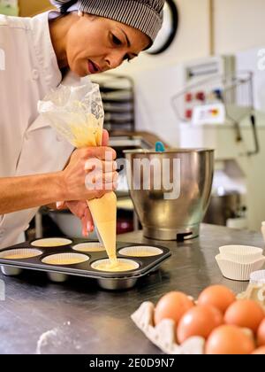 Professional female confectioner using pastry bag and filling baking form for muffins with dough while working in kitchen Stock Photo