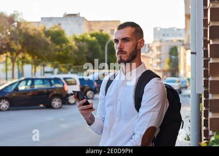 Serious adult bearded male in trendy ripped jeans with backpack checking information on mobile phone while walking with bicycle on paved urban street Stock Photo
