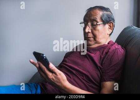 Senior ethnic male in glasses relaxing on couch in living room and reading messages on smartphone Stock Photo
