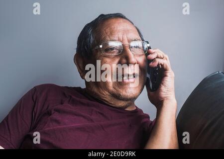 Elderly male sitting on sofa at home and speaking on mobile phone while looking away Stock Photo