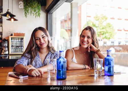 Smiling young girlfriends looking at camera while sitting at table with drinks during meeting in modern cafeteria in summer day Stock Photo
