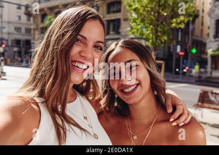 Cheerful young female friends embracing happily while taking selfie on urban square in summer day Stock Photo