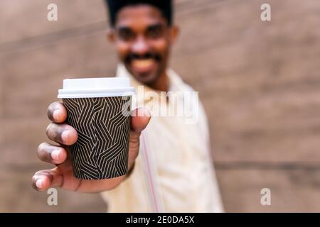 Blurred smiling black male showing disposable cup of takeaway coffee in outstretched hand and looking at camera friendly Stock Photo