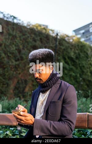 Serious African American male entrepreneur sitting on bench in urban park and writing plans in notebook Stock Photo