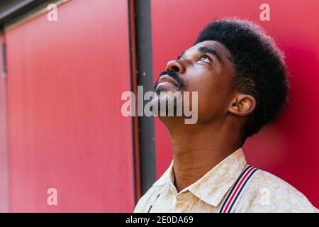 Tranquil African American male with curly hair leaning on red wall of building and looking up while dreaming Stock Photo