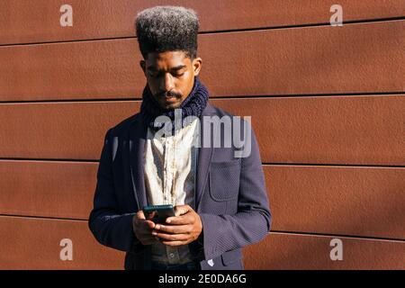 Focused African American male entrepreneur in stylish outfit standing near urban building and reading messages on mobile phone while working remotely Stock Photo