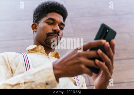 From below of serious bearded African American businessman in stylish casual shirt messaging on smartphone while standing against blurred wall of mode Stock Photo