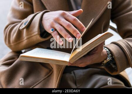 Cropped unrecognizable male in stylish outfit sitting outdoors reading interesting story in book Stock Photo