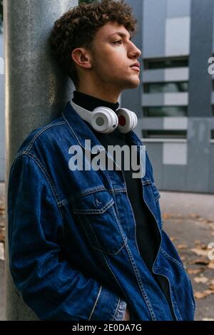 Thoughtful young male with curly hair wearing denim clothes with wireless headphones around the neck standing in city looking away Stock Photo