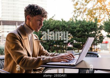 Side view of focused male freelancer sitting at table in outdoors cafe and typing on netbook while working remotely on project Stock Photo