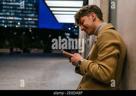 Side view of smiling handsome male leaning on building and messaging on social media via smartphone in evening in city Stock Photo