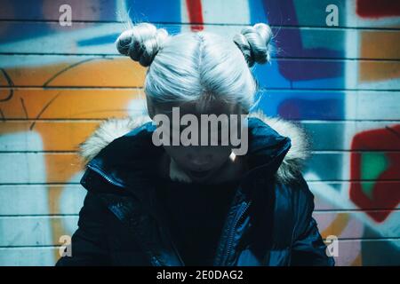 Asian teen hipster female with dyed hair wearing warm jacket looking down while standing under illumination against colorful wall with graffiti Stock Photo
