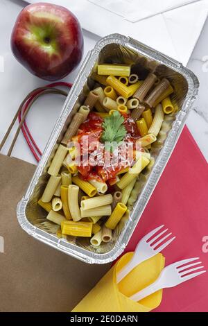Top view of foil box with delicious pasta with tomato sauce placed near fresh red apple on table with disposable forks and paper bags representing hea Stock Photo