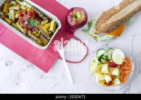 Top view composition with delicious pasta in foil box placed near salad bowl, fresh apple and sandwich with cheese and vegetables on table with paper Stock Photo