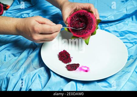 Crop anonymous person with spoon eating delicious dragon fruit placed on plate on blue background in studio Stock Photo