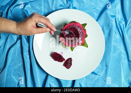 Top view of crop anonymous person with spoon eating delicious dragon fruit placed on plate on blue background in studio Stock Photo