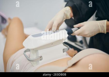 Cropped unrecognizable slim female client lying on table and getting anti cellulite massage of belly with LPG machine while looking at professional ma Stock Photo