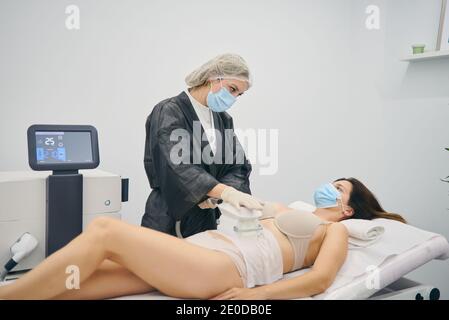 Slim female client lying on table and getting anti cellulite massage of belly with LPG machine while looking at professional masseuses Stock Photo