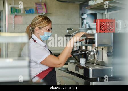 Side view of female barista in medical mask using coffee machine and filling cup with hot espresso while working in cafeteria during coronavirus pande Stock Photo