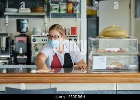 Female bartender in medical mask wiping counter with antibacterial nap while working in cafeteria during coronavirus pandemic Stock Photo