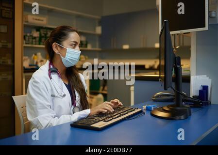 Side view of young female medic in white coat and protective mask typing on computer keyboard while working in cabinet of modern clinic Stock Photo