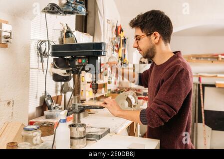 Side view of male woodworker using drilling tool while working with wood in bright carpentry workshop Stock Photo