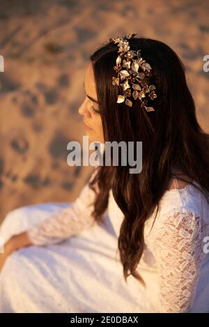 From above side view of young romantic bohemian brunette in white wedding dress and golden floral wreath sitting on sand Stock Photo