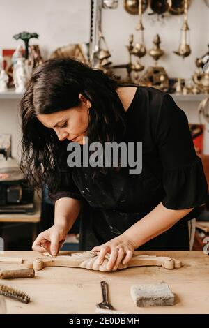 Skilled female artisan polishing decorative carved wooden molding with abrasive paper while working in professional workshop Stock Photo