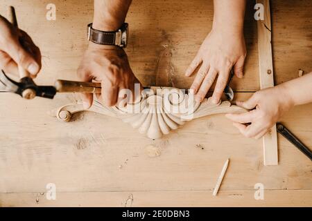 Top view of crop anonymous artisan masters with hammer and chisel carving decorative wooden detail while working together in workshop Stock Photo