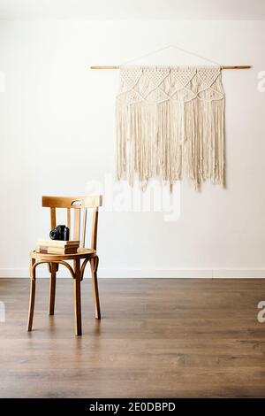 Minimalist interior of room with photo camera and books stacked on wooden stool placed against white wall with handmade boho hanging decoration Stock Photo