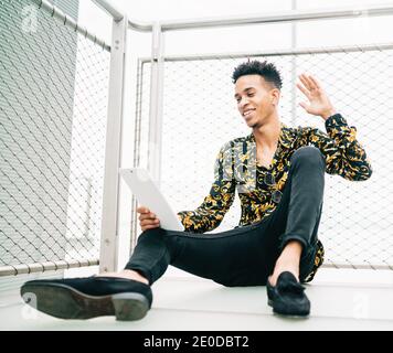 Cheerful African American male sitting on terrace with tablet and having video call while smiling and waving hand Stock Photo