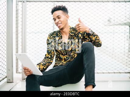 Cheerful African American male sitting on terrace with tablet and having video call while smiling and waving hand Stock Photo