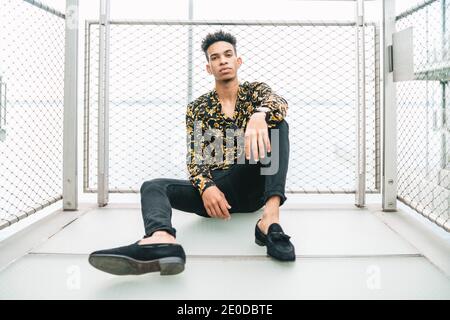 Handsome African American male in trendy apparel sitting on promenade near fence and looking at camera Stock Photo