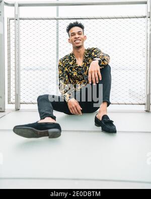 Handsome African American male in trendy apparel sitting on promenade near fence and looking at camera Stock Photo
