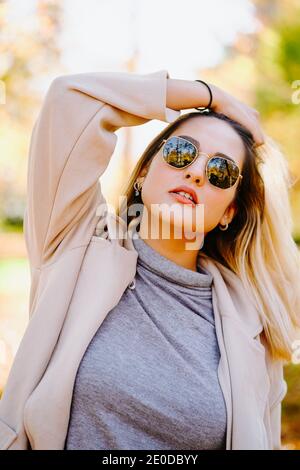 Smiling female wearing autumn coat and sunglasses standing in park on sunny day and looking at camera Stock Photo