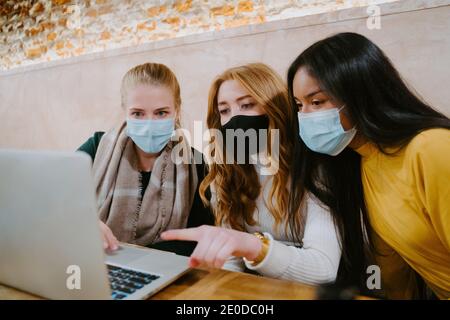 Group of multiethnic female freelancers in masks working on laptop in cafe while discussing new business project during coronavirus epidemic Stock Photo